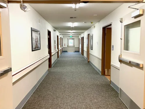 A long, well-lit hallway with carpeted floor, framed images on the walls, and multiple doors leading to rooms