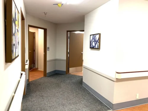 A well-lit hallway featuring textured carpet, wall art, and open doorways leading to adjacent rooms.